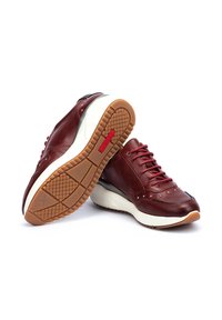 Un par de zapatillas de cuero rojo oscuro con medias suelas blancas y suelas exteriores de goma texturizada marrón, una zapatilla descansando de lado mostrando el patrón de la suela.