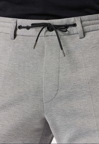 Grijze sweatpants van een gestructureerde stof, met een elastische tailleband, zwarte trekkoord en schuin geplaatste voorzakken. Metalen uiteinden op de trekkoord.
