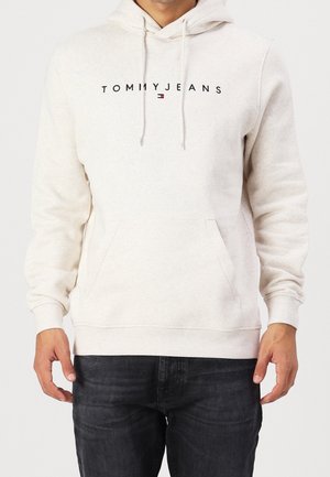 Mann trägt einen cremefarbenen Hoodie mit "TOMMY JEANS" Schriftzug und Logo, kombiniert mit dunklen Jeans, Hände entspannt an den Seiten.