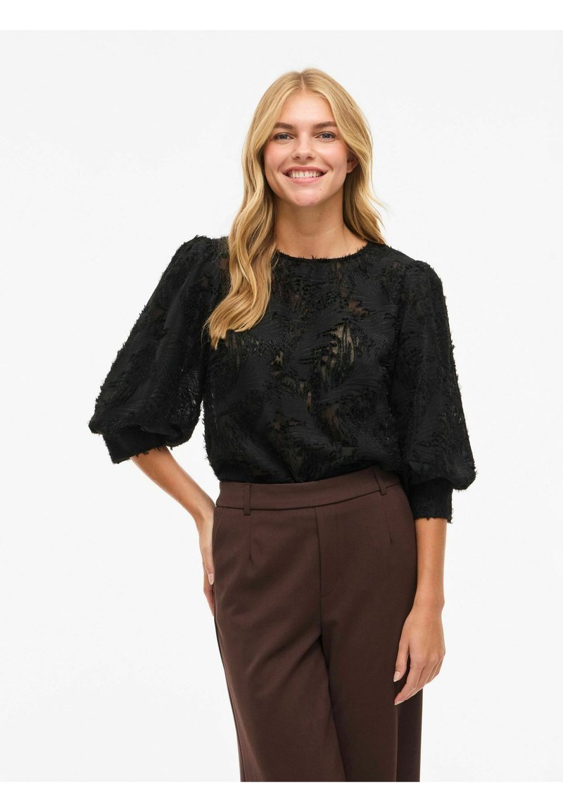 Svart bluse med teksturert stoff, ballongsleeves, rund halslinning og gjennomsiktig detaljer, kombinert med brune skreddersydde bukser.