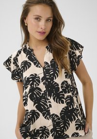 Blusa nera e crema con maniche corte, caratterizzata da una stampa di monstera fogliosa, colletto e una leggera forma ad A. Tessuto dalla texture liscia.