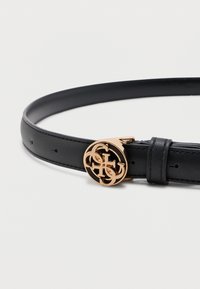 Μαύρη δερμάτινη ζώνη με χρυσή κυκλική αγκράφα που διαθέτει πλεγμένα διακοσμητικά γράμματα σε απλό λευκό φόντο.