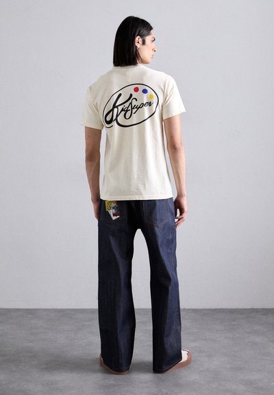 Cream short-sleeve t-shirt with black "KidSuper" logo on back. Worn with dark blue jeans featuring patch on left pocket. Flat, textured surface.