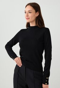 Czarny dzianinowy sweter z wysokim dekoltem i długimi rękawami. Posiada złote guziki na mankietach oraz ribbrowany dół dla dodatkowej tekstury.