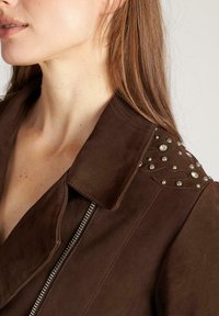 Brązowa skórzana kurtka z wyprofilowanym kołnierzem, srebrnymi ćwiekami na ramionach oraz zapięciem na zamek. Posiada fakturowaną powierzchnię oraz dopasowany krój.