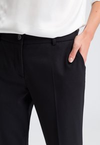 Schwarze Hosen mit glatter Textur, ausgestattet mit einem Knopfverschluss und seitlichen Taschen. Sichtbare Bügelfalte entlang der Vorderseite.