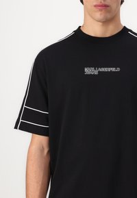 Črna bombažna majica s kratkimi rokavi in belimi poudarnimi črtami na ramenih ter belim logotipom "KARL LAGERFELD JEANS" na prsih. Ovratnik je okrogel.