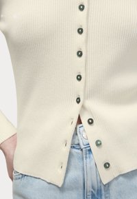 Cardigan crema a coste con bottoni verdi, caratterizzato da una vestibilità dritta, orlo spaccato e una texture sovrapposta su jeans di un blu chiaro.