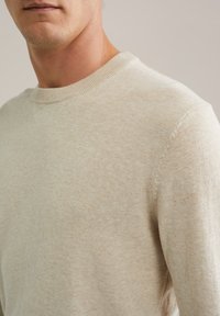 Primo piano di un uomo che indossa un maglione a girocollo lavorato a maglia beige, con dettagli sulla trama e sulle cuciture nelle aree delle spalle e del collo.