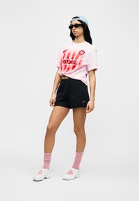 Ροζ γραφικό t-shirt με κόκκινο κείμενο, μαύρο σορτς, ροζ κάλτσες και λευκά αθλητικά παπούτσια. Αξεσουάρ περιλαμβάνουν ανοιχτό μπλε καπέλο και γυαλιά ηλίου.