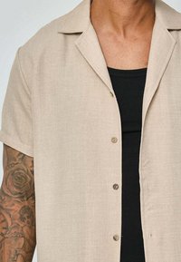Chemise beige à manches courtes en tissu texturé avec un col cranté et des boutons marron, superposée à un débardeur noir côtelé.