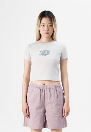 Ung kvinne med rett blondt hår iført en hvit kort t-skjorte med blomstertekst og teksturerte lyse lilla shorts, stående foran bakgrunn i hvitt.