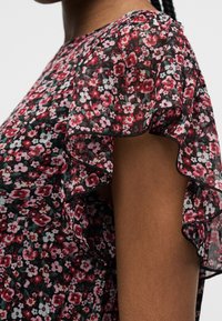 Gros plan d'un chemisier transparent à fleurs rouges, roses et blanches, avec des manches à volants superposés sur un fond de tissu sombre.