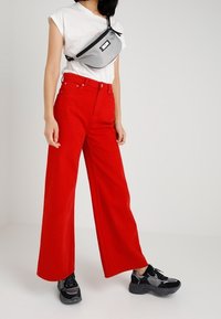 Czerwone szerokie spodnie zestawione z białą koszulką z krótkim rękawem i szarym torbą na pasku. Czarne sneakersy z teksturowanymi akcentami dopełniają wygląd.