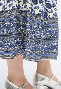 Μπλε και λευκή φούστα με λουλουδένιο σχέδιο, φορεμένη με ασημένια παπούτσια στιλ Mary Jane με χαμηλό τακούνι, σε γυμνά πόδια.