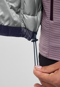 Doudoune argentée avec un ourlet bleu marine, dotée de cordons de serrage réglables. Tissu violet doux et texturé en dessous, avec un détail de poche zippée.
