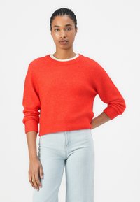Rode cropped trui van zacht breigoed, met geribbelde boorden en zoom, gecombineerd met lichtblauwe high-waisted jeans. Witte onderhemd zichtbaar.