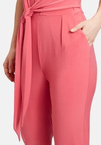 Rosa jumpsuit med et struktureret stof. Har en knyttet taljedetalje, plisseringer og sidelommer for funktionalitet.