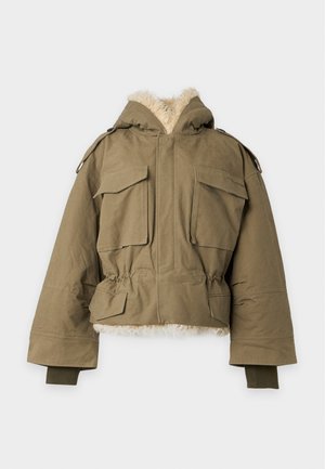 Mulher a usar um casaco oversized verde-oliva forrado a pelo, hoodie cinzento com texto vermelho e azul, saia patchwork de ganga e botas pretas até ao tornozelo.