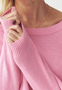 Růžový pletený svetr s širokým výstřihem, žebrovanými manžetami a měkkou texturou. Detailní pohled na ruce držící látku.