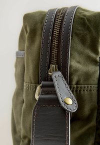 Olivgrön kanvasväska med en brun läderdragkedja med guldfärgad hårdvara, samt ett läderhandtag med kontrasterande sömdetaljer.