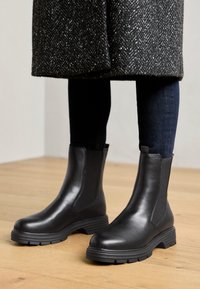 Μαύρες ankle μπότες με λεία επιφάνεια, ελαστικές πλαϊνές πάνελ, υφασμάτινη σόλα από καουτσούκ και χαμηλό τακούνι τύπου μπλοκ, συνδυασμένες με σκούρα τζιν.