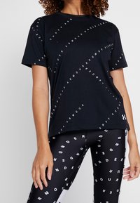 Svart t-shirt med vit text "UNDER ARMOUR" tryckt i diagonala mönster. Korta ärmar, avslappnad passform, tillverkad av mjukt tyg.