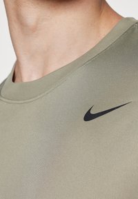 Tricou sportiv Nike de culoare verde măslinie, cu guler rotund, mâneci scurte, textură netedă și un logo swoosh negru pe piept.