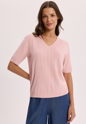 Kobieta nosząca jasnoróżowy sweter z krótkim rękawem i dekoltem w serek z subtelnym wzorem oraz niebieskie spodnie, stojąca na tle jednolitego tła.