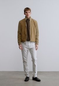 Καφέ σακάκι από καστόρι με φερμουάρ μπροστά, συνδυασμένο με πράσινο πλεκτό πουλόβερ με υφή και λευκά τζιν. Μαύρα παπούτσια ολοκληρώνουν το ντύσιμο.