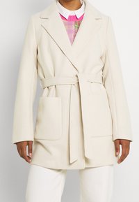 Cappotto beige chiaro con un design avvolgente, vita con cintura, rever a gola, e due tasche frontali, realizzato in un tessuto morbido.