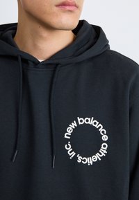Sweatshirt à capuche noir avec un logo circulaire blanc portant l'inscription "new balance athletics, inc." Le texte est positionné le long du bord du cercle.
