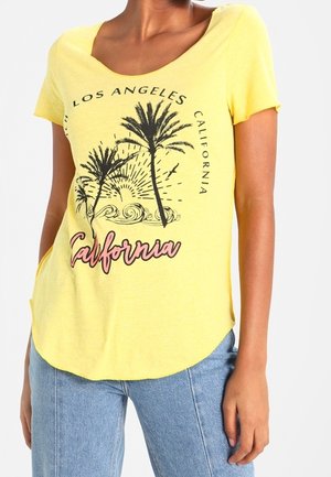 Femme portant un t-shirt jaune avec des palmiers, le soleil, des vagues, et le texte disant Los Angeles California et California en écriture cursive.