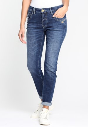 Vrouw draagt opgerolde donkerblauwe spijkerbroek met knoop en rits, witte sneakers en een wit topje, één hand in de zak.