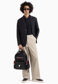 Zaino in pelle nera con accenti rossi, texture morbida, multiple tasche e dettagli con zip. Indossato con un blazer nero e pantaloni beige.