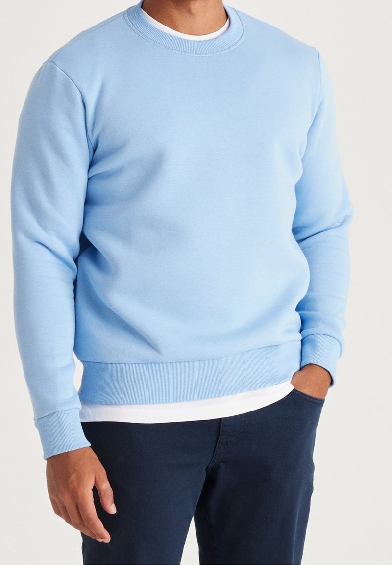 Homme portant un sweat-shirt à col rond bleu clair par-dessus une chemise blanche, associé à un pantalon bleu foncé, debout avec une main dans la poche.