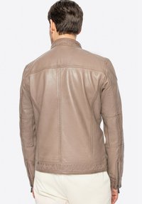 Giacca di pelle marrone con un design strutturato, caratterizzata da cuciture visibili, colletto alto e polsini con zip. Texture liscia con vestibilità su misura.