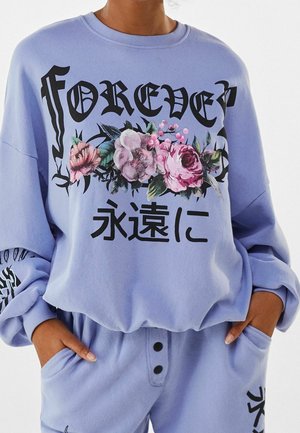 Komplet z bluzy i spodni w kolorze lawendowym z rękami w kieszeniach; bluza ozdobiona napisem "FOREVER", kwiatowym wzorem i czarnymi azjatyckimi znakami.
