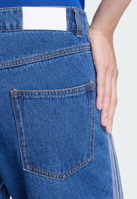 Μπλε τζιν παντελόνι με πίσω τσέπη που έχει πορτοκαλί ραφές και λευκή ετικέτα στη μέση. Ένα χέρι ακουμπά στην περιοχή του γοφού.