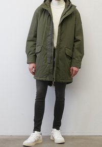 Olivgrön parka med huva, framdragkedja och tryckknappsstängning, med två stora framfickor och en lätt textur.