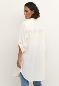 Tunique en lin blanc avec une coupe décontractée, des manches retroussées et un détail de patte de boutonnage au dos. L'ourlet est arrondi et partiellement fendu sur les côtés.