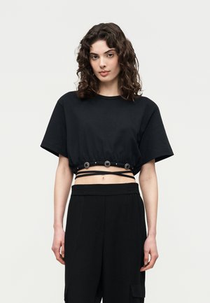 Fiatal nő göndör hajjal, fekete hímzett gombos, körbetekerős pántos rövid felsőben, fekete nadrággal párosítva.