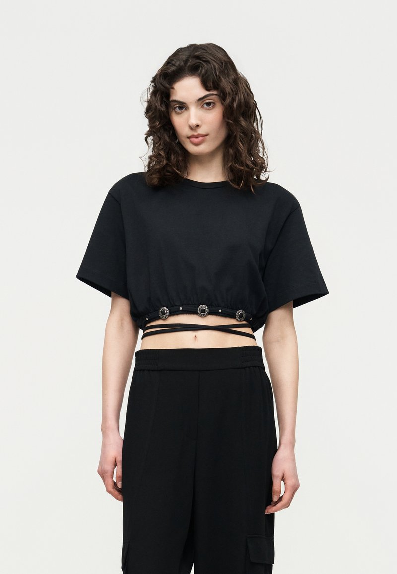 Νέα γυναίκα με μαλλιά σε μπούκλες, που φοράει ένα μαύρο κοντό τοπ με διακοσμητικά κουμπιά και λουριά που τυλίγονται γύρω, συνδυασμένο με μαύρο παντελόνι.