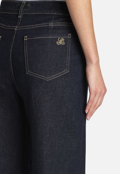 Mørke denimjeans med en tætsiddende pasform og synlige sømme. Baglommen har et diskret broderet logo i guld.