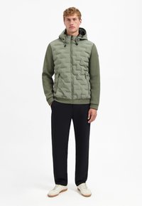 Olijfgroene jas met een gewatteerde bovenkant, capuchon en geribde mouwen. Gecombineerd met zwarte broeken en witte sneakers. Eenvoudig ontwerp, functionele stijl.