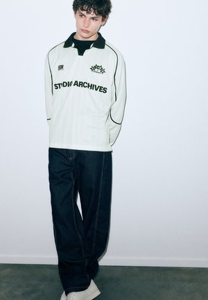 Langärmeliges, helles Hemd mit vertikalen Streifen, schwarzem Kragen und dem Text "STUDIO ARCHIVES". Dunkelblaue Jeans mit lockerem Schnitt. Weiße Turnschuhe.