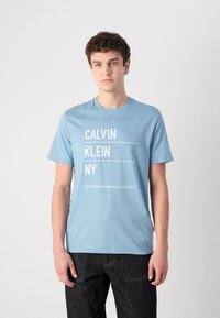 Šviesiai mėlynas medvilninis marškinėliai, su baltais spausdintais tekstais "CALVIN KLEIN NY" ir "Calvin Klein Established 1968 NYC". Apvalus kaklas, trumpomis rankovėmis.