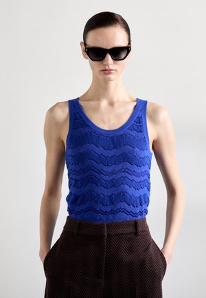 Top in maglia blu testurizzato con un motivo ondulato, abbinato a pantaloni marrone a spina di pesce. Il modello indossa occhiali da sole neri.