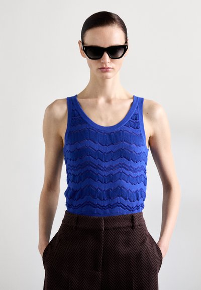 Modrá texturovaná pletená tílko s vlnkovým vzorem, spárované s hnědými herringbone kalhotami. Modelka nosí černé sluneční brýle.