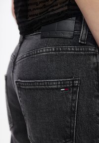 Κοντινό πλάνο μαύρου τζιν με ορατή πίσω τσέπη, δερμάτινο έμπλαστρο με την επιγραφή "JEANS" και μικρό υφασμάτινο λογότυπο σε κόκκινο, λευκό και μπλε χρώμα.